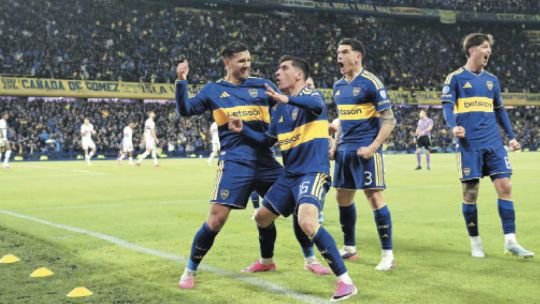 Jugadores de fútbol disputando la pelota en un partido, con un jugador dejando pasar el balón entre sus piernas.